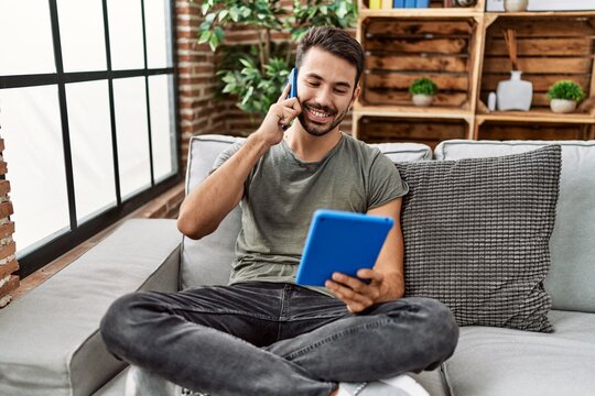 Young hispanic man using touchpad and talking on the smartphone at home