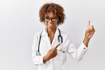 Young african american woman wearing doctor uniform and stethoscope smiling and looking at the camera pointing with two hands and fingers to the side.