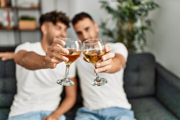 Two hispanic men couple toasting with glass of wine sitting on sofa at home