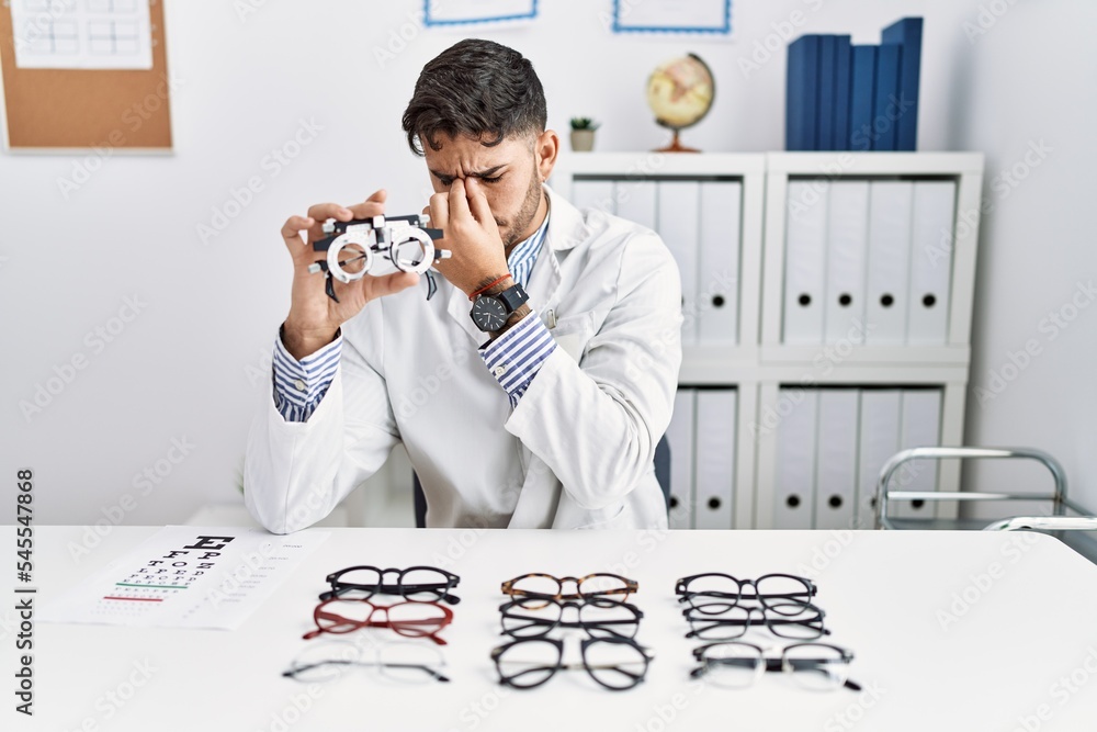 Sticker Young optician man holding optometry glasses tired rubbing nose and eyes feeling fatigue and headache. stress and frustration concept.