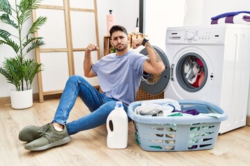 Young hispanic man putting dirty laundry into washing machine showing arms muscles smiling proud....