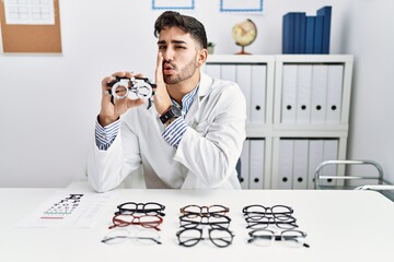 Young optician man holding optometry glasses hand on mouth telling secret rumor, whispering...