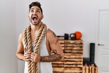 Young hispanic man training with battle rope at the gym angry and mad screaming frustrated and furious, shouting with anger looking up.