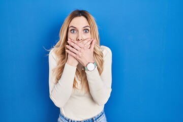Young caucasian woman standing over blue background shocked covering mouth with hands for mistake....