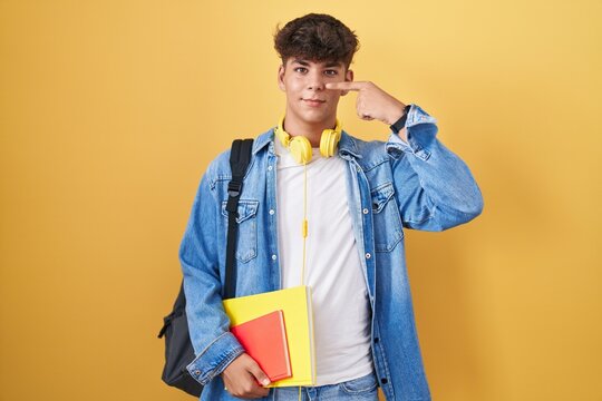 Hispanic teenager wearing student backpack and holding books pointing with hand finger to face and nose, smiling cheerful. beauty concept