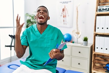 Fototapeta premium Young african american man working at pain recovery clinic crazy and mad shouting and yelling with aggressive expression and arms raised. frustration concept.