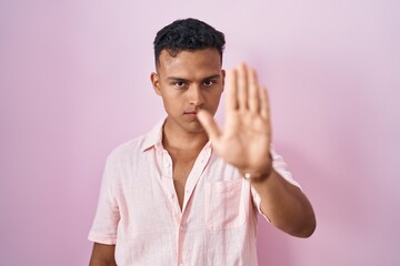 Young hispanic man standing over pink background doing stop sing with palm of the hand. warning...