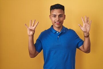 Young hispanic man standing over yellow background showing and pointing up with fingers number nine...