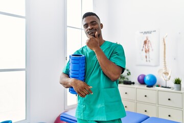 Young african physiotherapist man holding foam roller at the clinic serious face thinking about...