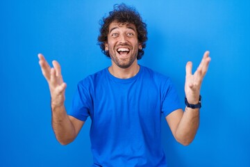 Naklejka premium Hispanic young man standing over blue background celebrating crazy and amazed for success with arms raised and open eyes screaming excited. winner concept