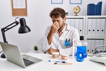 Young hispanic dentist man working at medical clinic tired rubbing nose and eyes feeling fatigue...