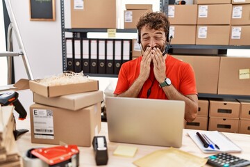 Young hispanic call center agent man working at warehouse laughing and embarrassed giggle covering...