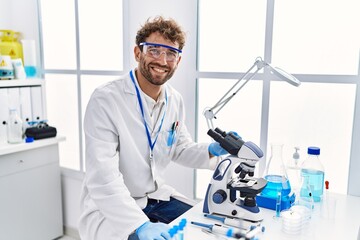 Young hispanic man wearing scientist uniform using microscope at laboratory