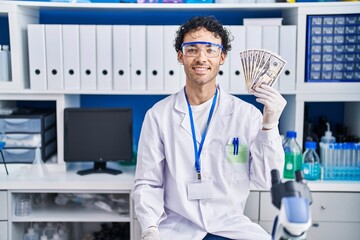 Hispanic man working at scientist laboratory holding money looking positive and happy standing and smiling with a confident smile showing teeth