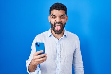 Hispanic man with beard using smartphone typing message sticking tongue out happy with funny expression. emotion concept.