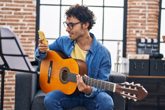 Young hispanic man musician playing classical guitar using smartphone at music studio