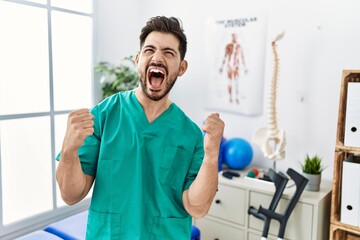 Young man with beard working at pain recovery clinic very happy and excited doing winner gesture with arms raised, smiling and screaming for success. celebration concept.