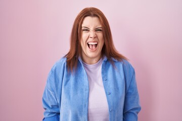 Young hispanic woman with red hair standing over pink background angry and mad screaming frustrated...