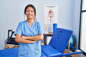 Young chinese woman wearing physiotherapist uniform standing with arms crossed gesture at rehab...