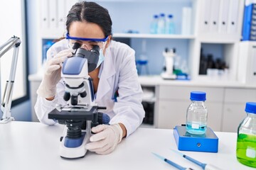 Young hispanic woman wearing scientist uniform using microscope at laboratory