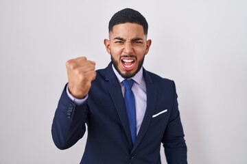 Young hispanic man wearing business suit and tie angry and mad raising fist frustrated and furious while shouting with anger. rage and aggressive concept.