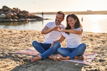 Middle age man and woman couple doing heart symbol with hands sitting on sand at seaside