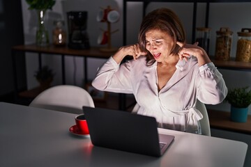 Middle age hispanic woman using laptop at home at night covering ears with fingers with annoyed expression for the noise of loud music. deaf concept.