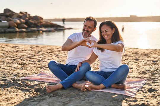 Middle age man and woman couple doing heart symbol with hands sitting on sand at seaside