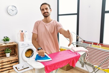 Young hispanic man ironing clothes at home smiling friendly offering handshake as greeting and welcoming. successful business.