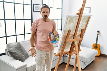 Young hispanic man with beard painting on canvas at home in shock face, looking skeptical and sarcastic, surprised with open mouth