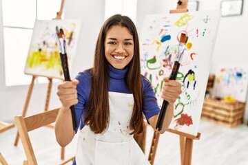 Young latin woman smiling confident holding paintbrushes at art studio