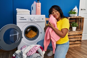 Young beautiful latin woman washing clothes touching face with soft sweater at laundry room