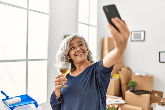 Middle age grey-haired woman toasting with wine making selfie by the smartphone at new home.
