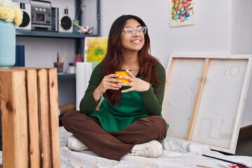Young hispanic woman artist drinking coffee sitting on floor at art studio