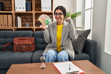 Young hispanic woman working at consultation office holding money surprised with an idea or...