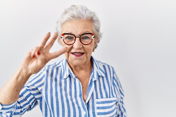 Senior woman with grey hair standing over white background showing and pointing up with fingers number three while smiling confident and happy.