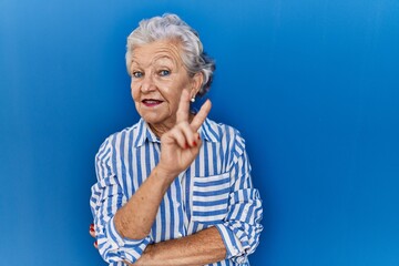 Senior woman with grey hair standing over blue background smiling with happy face winking at the...