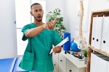 African american physiotherapist man working at pain recovery clinic pointing with finger surprised ahead, open mouth amazed expression, something on the front