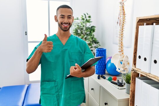 African american physiotherapist man working at pain recovery clinic doing happy thumbs up gesture with hand. approving expression looking at the camera showing success.