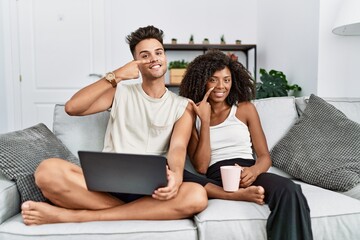 Young interracial couple using laptop at home sitting on the sofa pointing with hand finger to face and nose, smiling cheerful. beauty concept