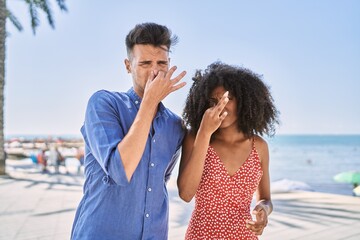 Young interracial couple outdoors on a sunny day smelling something stinky and disgusting, intolerable smell, holding breath with fingers on nose. bad smell