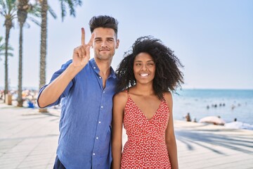 Young interracial couple outdoors on a sunny day showing and pointing up with fingers number two while smiling confident and happy.