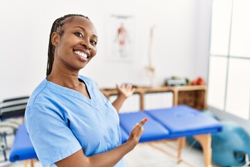 Black woman with braids working at pain recovery clinic inviting to enter smiling natural with open hand