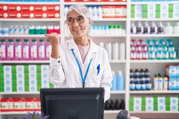 Middle age woman with tattoos working at pharmacy drugstore smiling with happy face looking and pointing to the side with thumb up.