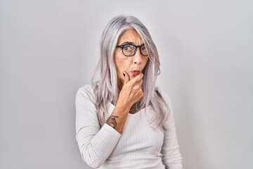 Middle age woman with grey hair standing over white background looking fascinated with disbelief, surprise and amazed expression with hands on chin