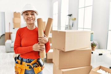 Middle age grey-haired woman smiling confident holding blueprints at new home