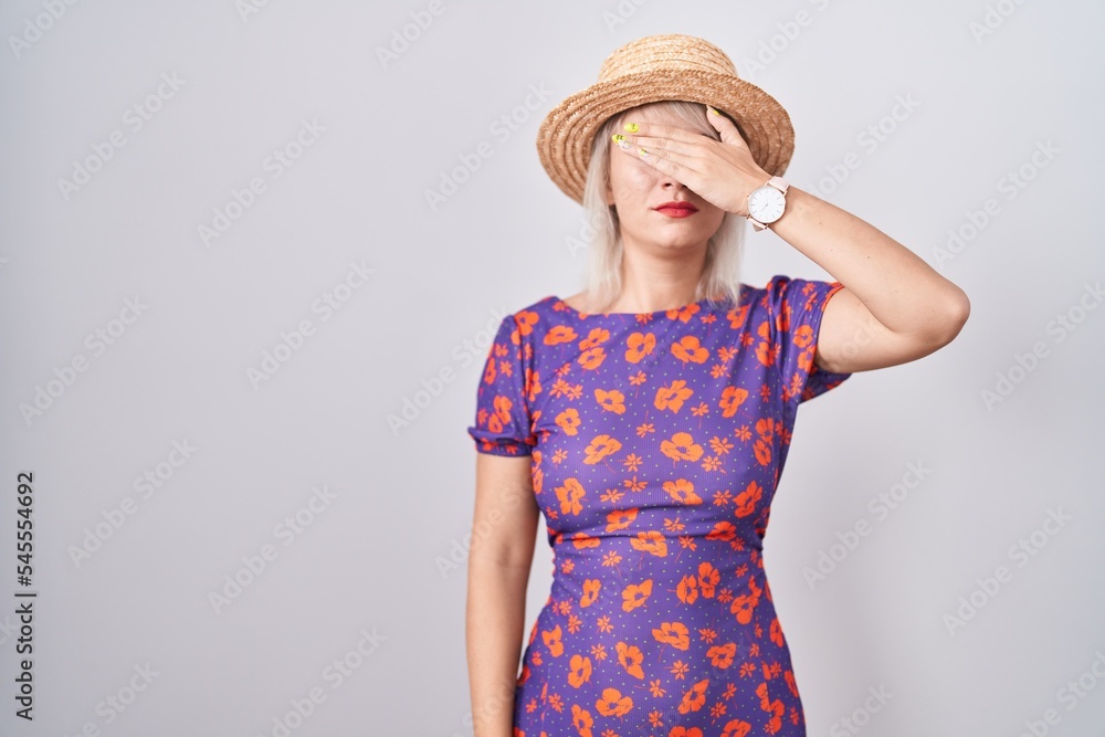 Sticker Young caucasian woman wearing flowers dress and summer hat covering eyes with hand, looking serious and sad. sightless, hiding and rejection concept