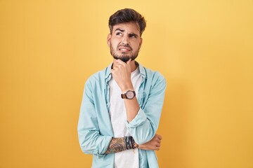 Fototapeta premium Young hispanic man with tattoos standing over yellow background thinking worried about a question, concerned and nervous with hand on chin