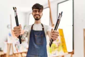Young hispanic artist man smiling happy holding paintbrushes at art studio.