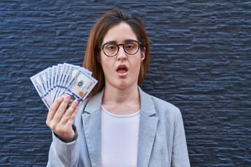 Brunette woman holding dollars scared and amazed with open mouth for surprise, disbelief face